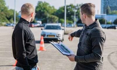A driving instructor explain rules to a learner driving at a test centre with a car far at the distant background